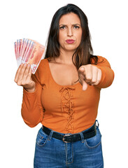Beautiful hispanic woman holding norwegian krone banknotes pointing with finger to the camera and...