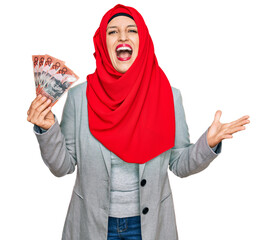 Beautiful hispanic woman wearing islamic hijab holding australian dollars banknotes celebrating victory with happy smile and winner expression with raised hands