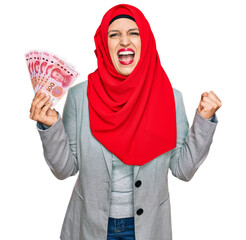 Beautiful hispanic woman wearing islamic hijab holding chinese yuan banknotes screaming proud, celebrating victory and success very excited with raised arms