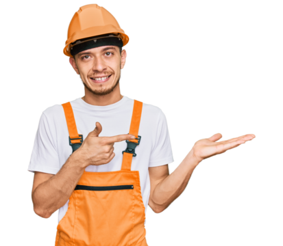 Hispanic young man wearing handyman uniform and safety hardhat amazed and smiling to the camera while presenting with hand and pointing with finger.