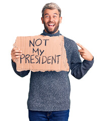 Young handsome blond man holding not my president cardboard banner smiling happy pointing with hand...