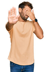Handsome young man with beard wearing casual tshirt covering eyes with hands and doing stop gesture...