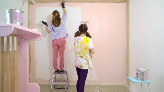 Back view of happy mother and daughter painting house wall in pink color using roller brush in apartment renovating home. House redesigning, renovating and repair concept.
