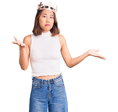 Young beautiful chinese girl wearing princess crown clueless and confused expression with arms and hands raised. doubt concept.