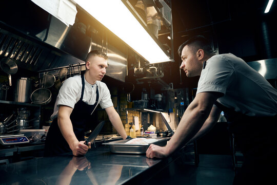 Competitor chefs with knives looking at each other at table in restaurant - Powered by Adobe
