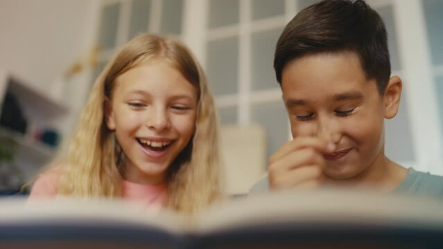 Young girl and boy reading a book together, laughing at funny pictures, leisure