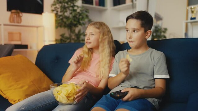 Two children friends eating chips and watching movie at home, leisure activity