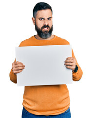 Hispanic man with beard holding blank empty banner clueless and confused expression. doubt concept.
