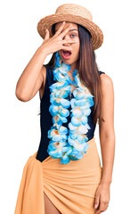 Young beautiful latin girl wearing hawaiian lei and summer hat peeking in shock covering face and...