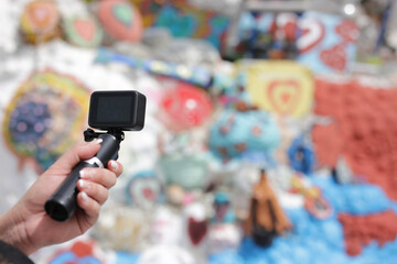 A person is holding a camera with a black body and a black grip. The camera is pointed at a wall with a lot of colorful graffiti. The wall is covered in various shapes and sizes of rocks