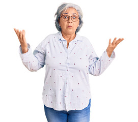 Senior woman with gray hair wearing casual business clothes and glasses clueless and confused expression with arms and hands raised. doubt concept.