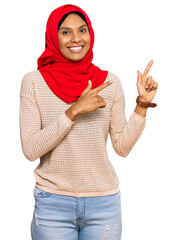 Young african american woman wearing traditional islamic hijab scarf smiling and looking at the camera pointing with two hands and fingers to the side.