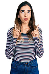 Beautiful hispanic woman wearing casual striped shirt amazed and surprised looking up and pointing with fingers and raised arms.