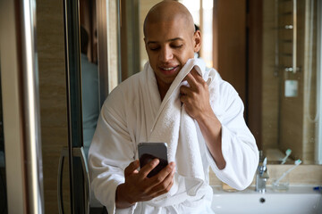 Young man drying face with towel and watching smartphone in bathroom