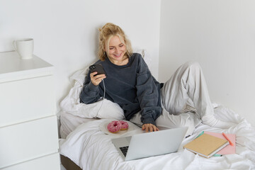 Portrait of smiling candid woman, lying in bed with doughnut, using smartphone and laptop, resting at home in bedroom, watching tv show or chatting online