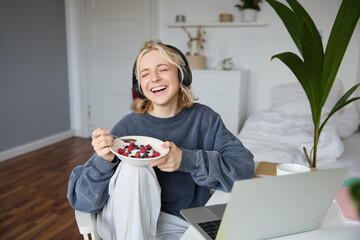 Portrait of happy young blond woman, sitting in a room, watching movie on laptop and eating healthy...