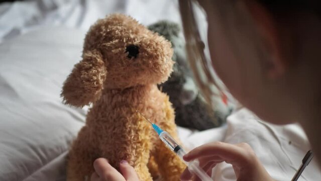 A little girl gives a vaccination injection to her stuffed dog using a syringe with a needle while playing veterinary hospital on her bed. A child treats a plush toy with an injection.