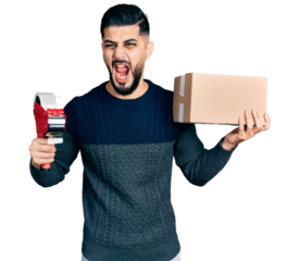 Young arab man with beard holding packing tape and cardboard box angry and mad screaming frustrated and furious, shouting with anger. rage and aggressive concept.