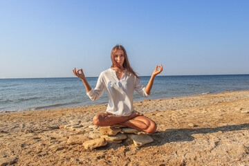 Fototapeta na wymiar A beautiful girl sits on a stone in the lotus position against the background of the sea and sky