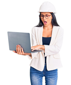 Young beautiful latin girl wearing architect hardhat holding laptop scared and amazed with open mouth for surprise, disbelief face
