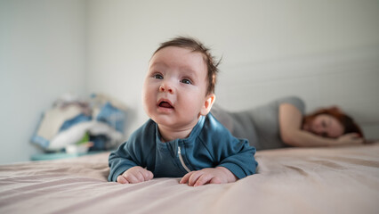 A three-month-old boy lies on his stomach on the bed and his mother sleeps behind him. Postpartum...
