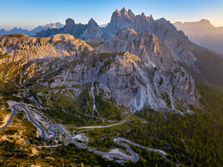 Drone panoramic shot of a windings road to Tre Cime di Lavaredo peaks in the Dolomites, sharp summits in the background lit by the setting sun in vibrant orange-blue tones. UNESCO site, Italy.