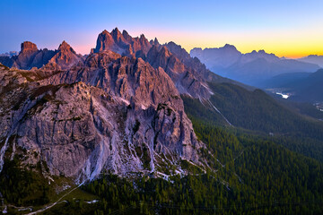 Aerial view from high altitude of Dolomite peaks lit by pink light of  the sunset sky, with Lago di Misurina valley and Dolomites mountain panorama against blue sky with a yellow horizon band. Italy