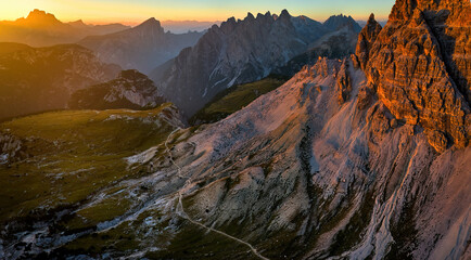 Drone panoramic shot of a hiking trail around Tre Cime di Lavaredo peaks in the Dolomites, with sharp summits in the background lit by the setting sun in vibrant orange-blue tones.