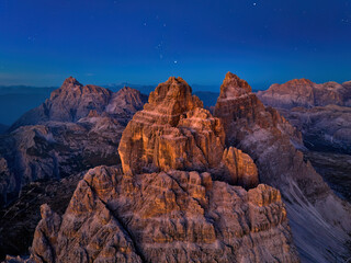 Golden-lit peaks of Tre Cime di Lavaredo from a high altitude, at rock summit level. The valley below is dark, with a dark blue sky and stars above, illuminating the Dolomites panorama. Blue-gold tone