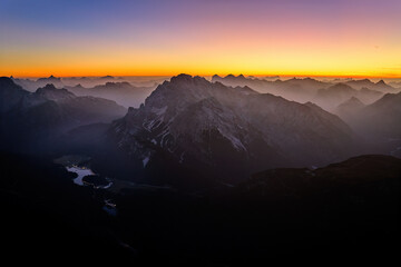 Dolomites peaks rise from valley fog at dawn, horizon blends from yellow-orange to dark blue. Drone aerial shot. Italy.