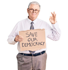 Senior grey-haired man wearing business clothes holding save our democracy protest banner doing ok sign with fingers, smiling friendly gesturing excellent symbol