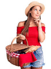 Young beautiful hispanic woman wearing summer hat and holding picnic wicker basket with bread serious face thinking about question with hand on chin, thoughtful about confusing idea