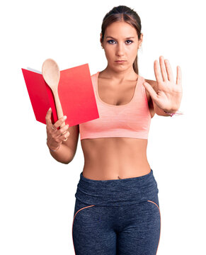 Young beautiful hispanic woman wearing gym clothes reading cooking recipe book holding spoon with open hand doing stop sign with serious and confident expression, defense gesture