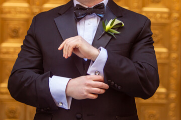 Groom in tuxedo and white calla lily boutonniere fastens his diamond cufflinks in front of a golden background - obrazy, fototapety, plakaty