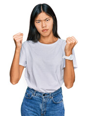 Beautiful young asian woman wearing casual white t shirt angry and mad raising fists frustrated and furious while shouting with anger. rage and aggressive concept.