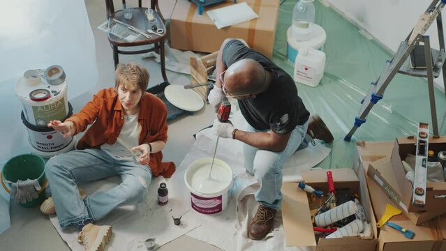 Young Caucasian woman sitting on the floor in room under renovation and sharing her thoughts about wall color with Black husband stirring paint in bucket with drill. High angle view