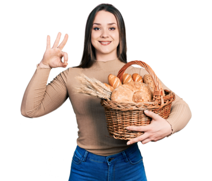 Young hispanic woman holding wicker basket with bread doing ok sign with fingers, smiling friendly gesturing excellent symbol