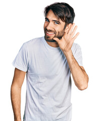 Hispanic man with blue eyes wearing casual white t shirt smiling with hand over ear listening and hearing to rumor or gossip. deafness concept.