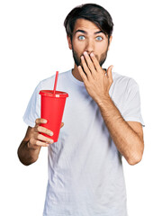 Hispanic man with blue eyes drinking glass of cola beverage covering mouth with hand, shocked and afraid for mistake. surprised expression