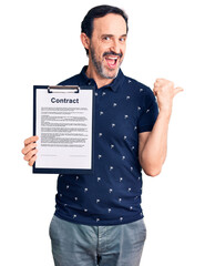 Middle age handsome man holding clipboard with contract document pointing thumb up to the side smiling happy with open mouth