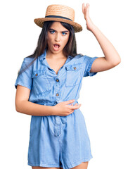 Brunette teenager girl wearing summer hat surprised with hand on head for mistake, remember error....