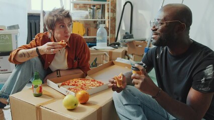 Young diverse couple sitting on the floor in living room, talking and eating pizza when having lunch break during home renovation - Powered by Adobe