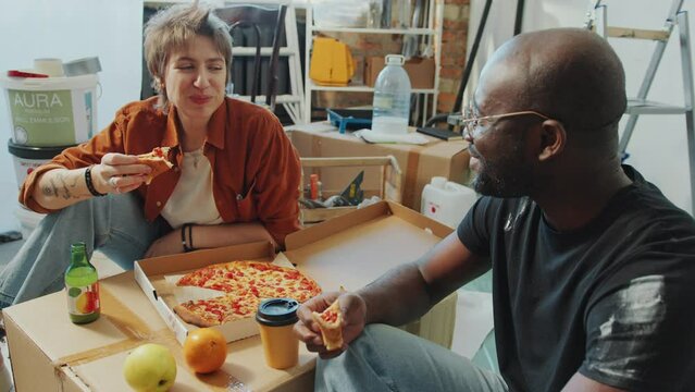 Young Caucasian woman and Black man sitting eating pizza, smiling and chatting when having lunch break during home renovation in room with ladder, paint and boxes