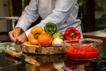 Chef lavando as mão ao fundo e na bancada a frente  ingredientes coloridos culinária saudável e uma faca e uma tigela de molho de tomate.