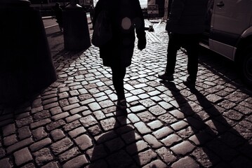 Silhouetted pedestrians walking on cobblestone street with shadows, urban setting.