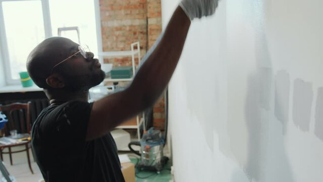Black man in gloves and stained t-shirt applying paint to the wall with roller in living room when doing home renovation