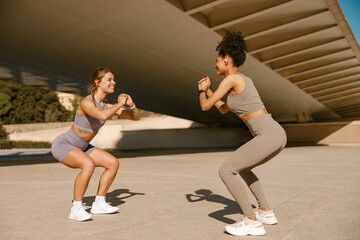Two smiling fit women doing workout and squatting together outdoors in the city