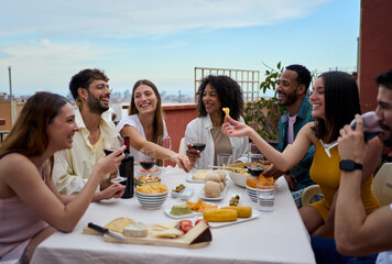 Laughing group of diverse young friends enjoying lunch together outdoors. Cheerful people gathered drinking red wine and eating snack on summer day having fun celebrating a birthday party on rooftop 