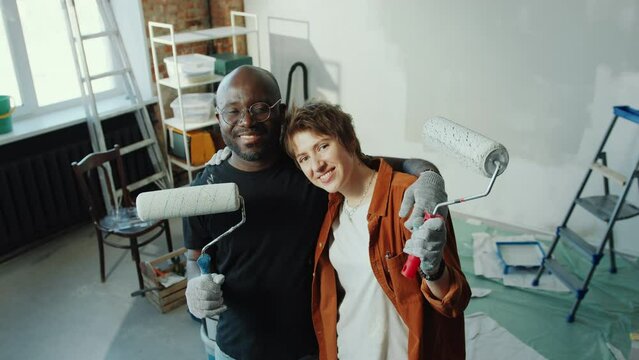 Joyous Caucasian wife and African American husband holding paint rollers, embracing and looking at camera with smile in home under renovation. High angle view, video portrait