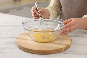 Woman whisking eggs in bowl at light marble table indoors, closeup